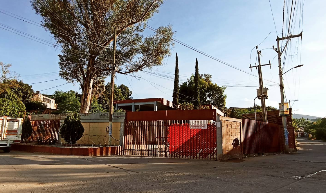 Fotografía en la que se muestra la entrada del plantel, conformada por una reja de color rojo y al lado izquierdo de la rej<a hay una jardinera.